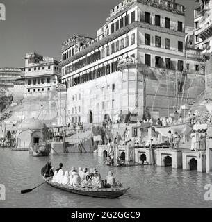 Historische indianer der 1950er Jahre, die in einem Ruderboot auf dem Ganges-Fluss in der Nähe des Flussufers mit seinen hohen Gebäuden in der Stadt Benares, Indien, sitzen. Als einer der größten Flüsse der Welt ist er als „heiliger Fluss“ bekannt, da die indianer, die Mehrheit der Hindus, ihn für heilig halten und seine Gewässer für religiöse Rituale nutzen, viele glauben, dass das Baden im Wasser des Ganges ihre Seelen reinigen wird. Der Ganges ist daher ein wichtiger Ort indischer religiöser Aktivitäten, Kultur und Geschichte. Stockfoto