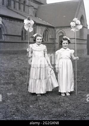 1956, historischer Karneval der May Queen, draußen auf dem Gelände einer Kirche, stehen zwei junge Mädchen für ein Foto in den Kostümen, die sie bei der Parade zum 1. Mai in der Stadt, England, Großbritannien, tragen werden. Sie halten die Blütenstöcke, die sie in der Prozession tragen werden. Stockfoto