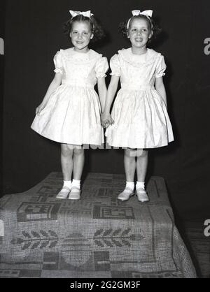 1956, historisch, May Queen Carnival, zwei junge Mädchen, Schwestern, Die Teilnahme an der traditionellen Parade zum 1. Mai steht zusammen auf einer Kiste mit Teppich und hält die Hände für ihr Foto in den hübschen Kleidern oder Outfits, die sie in der Prozession tragen werden, Leeds, England, Großbritannien. Stockfoto