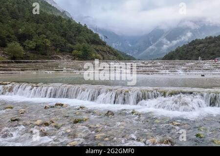 Kaskadierung von Mini-Wasserfällen des Baishui-Flusses, umgeben von den nebelbedeckten Gipfeln des Jade Dragon Snow Mountain im Blue Moon Valley, Yunnan, China Stockfoto