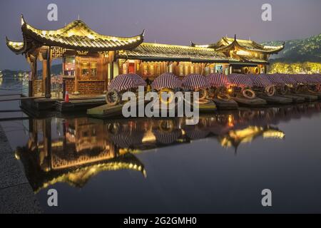 Verankerte Boote an einem beleuchteten Pier im chinesischen Design in West Lake bei Nacht mit Reflexionen im stillen Wasser, Hangzhou, Provinz Zhejiang, China Stockfoto