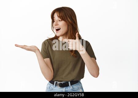 Erstaunt und glücklich junge Frau, zeigt und schaut auf ihre offene Hand, hält Objekt, zeigt Objekt auf der Handfläche gegen leere Kopie Raum, stehen über Stockfoto