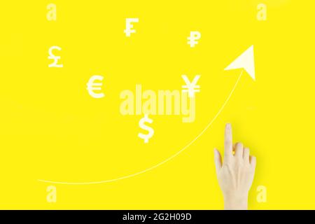 Konzept des globalen Währungswechsels. Die Hand einer jungen Frau zeigt mit dem Finger, den Währungen der Welt mit Hologramm und einem Pfeil nach oben auf gelbem Hintergrund. Erfolgreiche inte Stockfoto