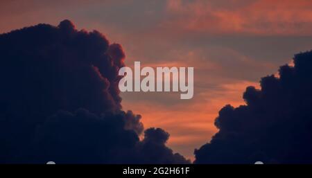 Dramatischer oranger Himmel zwischen dunklen dicken Wolken, die vom roten Sonnenuntergang beleuchtet werden und sich hinter den Wolken verstecken Stockfoto