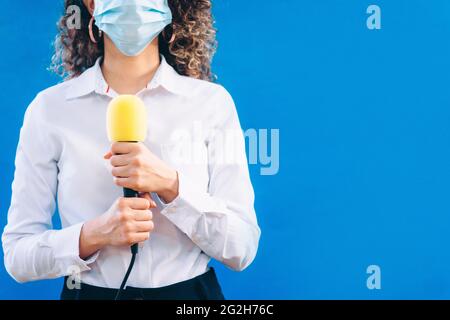 Reporterin trägt eine medizinische Gesichtsmaske mit einem gelben Mikrofon auf blauem Hintergrund Stockfoto