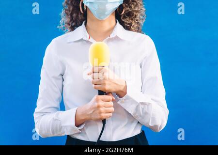 Reporterin trägt eine medizinische Gesichtsmaske mit einem gelben Mikrofon auf blauem Hintergrund Stockfoto