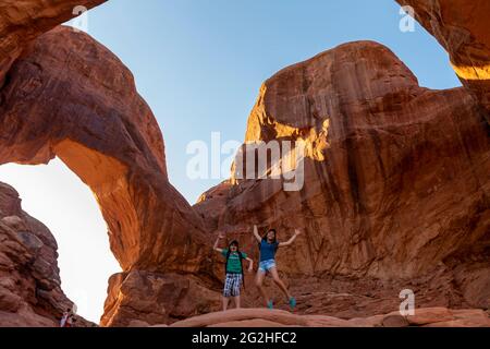 Zwei glückliche Kinder, die vor dem berühmten Double Arch springen - eine Sandsteinformation und beliebter Fotoport mit zwei großen Bögen, die aus dem gleichen Seitenfundament hervorgehen - bekannt für Vorder- und Rückspannen im Arches National Park, nahe Moab in Utah, USA Stockfoto