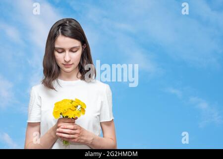 Hübsches kaukasisches Mädchen, das einen Strauß gelber Dandelionen vor einem blauen Himmel mit Wolken hält, Raum kopieren. Das Mädchen erhielt einen Strauß Wildblumen als Stockfoto