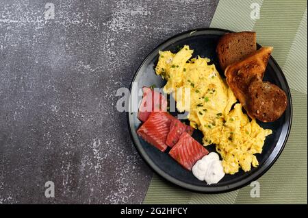 Rührei mit Lachs und Toast auf einem runden Teller auf dunklem Grund. Draufsicht, flach liegend Stockfoto