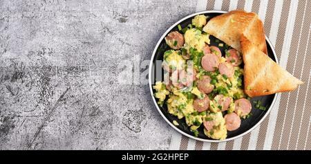 Rührei mit Wurst und Toast auf einem runden Teller auf dunklem Grund. Draufsicht, flach liegend Stockfoto