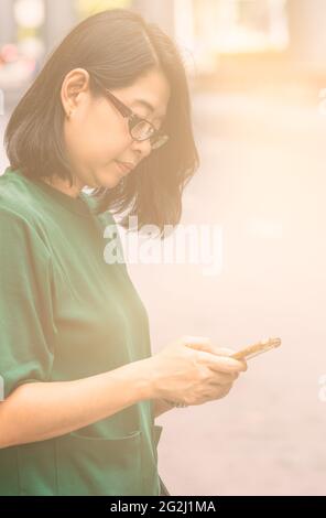 Porträt der schönen asiatischen Frau mittleren Alters SMS mit Smartphone auf der Straße Bürgersteig, Seitenansicht, vertikale Bildvorlage. Stockfoto