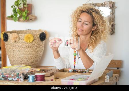 Erwachsene Frau, die handgemachte Kreationen Schmuck zu Hause mit bunten Perlen und Schnüren macht - weibliche Menschen, die zu Hause für Online-Shop Business-Shopping arbeiten - Handwerk diy Hobby weiblich und Juwelen auf einem Holztisch Stockfoto