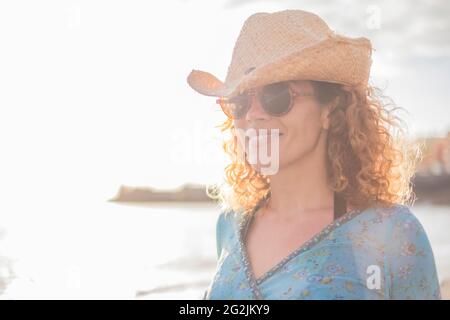 Porträt einer fröhlichen, glücklichen erwachsenen Frau im mittleren Alter im Freien, die lächelt und genießt - junge kaukasische Frauen mit Hut und Sonnenbrille - Meeresstrand im unfokussierten Hintergrund Stockfoto