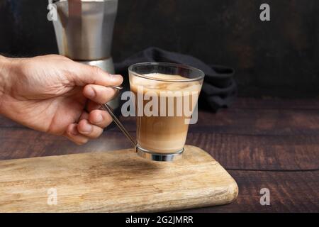 Die Hand des Mannes hält eine Tasse Kaffee mit Milch auf dem Holztisch und im Hintergrund eine italienische Kaffeekocher Stockfoto