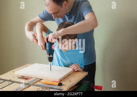 Vater und Sohn arbeiten zusammen, während sie an der Werkbank stehen. Kleiner Junge lernt zu bohren. Nettes Kind hält Bohrschrauber und Mann hilft ihm Stockfoto