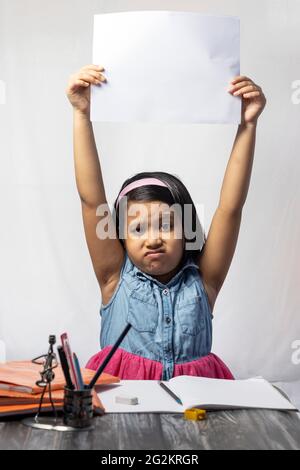 Ein unglückliches indisches Mädchen, das während des Studiums auf weißem Hintergrund ein leeres Blatt Papier über den Kopf hält Stockfoto