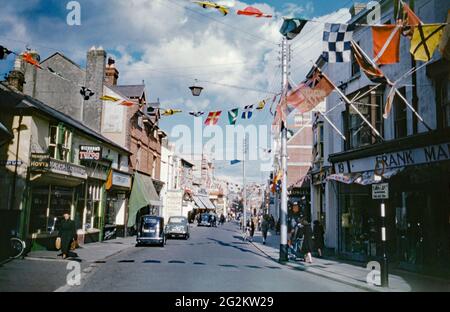 Auf der anderen Straßenseite gibt es farbenfrohe Fahnen und 1953 fliegen in der High Street, Littlehampton, West Sussex, England, Großbritannien, Gewerkschaftsflaggen. Dies war für die Krönung von Elizabeth II., die am 2. Juni 1953 in Westminster Abbey, London, stattfand. Elisabeth II. Trat im Alter von 25 Jahren nach dem Tod ihres Vaters, Georg VI., 1952 den Thron an. Auf der Straße befand sich das Art Deco Odeon Kino (Mitte links). Diese wurde abgerissen und die Straße ist jetzt autofrei. . Dieses Bild stammt von einem alten 35-mm-Kodak-Amateurfoto, einem Vintage-Foto aus den 1950er Jahren. Stockfoto