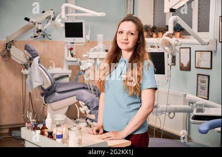 Schöne junge Frau Zahnarzt Blick auf Kamera und lächelnd, während am Tisch mit Instrumenten und Werkzeugen in der Zahnarztpraxis stehen. Konzept der Zahnmedizin und Zahnpersonal. Stockfoto