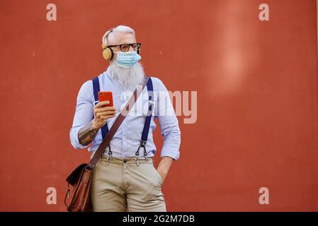 Stilvoller Seniorchef mit Headset und steriler Maske mit Smartphone Stockfoto
