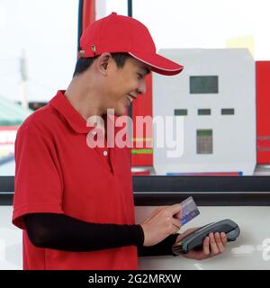 Tankstellenmitarbeiter in roter Uniform stehen lächelnd und wischen über das Zahlungsterminal mit einer Mockup-Kreditkarte. Anzeige von Bargeld, Preis und Volumen an der Zapfsäule Stockfoto