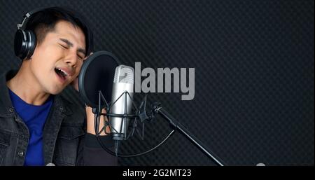 Junger asiatischer Mann mit Kopfhörern, der vor einer schwarzen Schallschutzwand singt. Musiker, der Musik im professionellen Aufnahmestudio produziert. Stockfoto