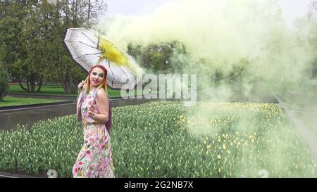 Entzückendes Mädchen in einem Kleid mit einem Blumendruck mit Regenbogen Zöpfen und Make-up. Tanzt mit einem Regenschirm, der sich in gelbem Rauch vor der Kulisse von FL Stockfoto