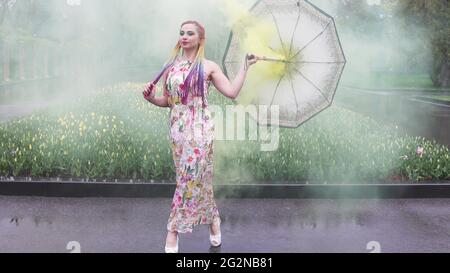 Entzückendes Mädchen in einem Kleid mit einem Blumendruck mit Regenbogen Zöpfen und Make-up. Tanzt mit einem Regenschirm, der sich in gelbem Rauch vor der Kulisse von FL Stockfoto