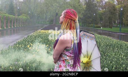 Entzückendes Mädchen in einem Kleid mit einem Blumendruck mit Regenbogen Zöpfen und Make-up. Tanzt mit einem Regenschirm, der sich in gelbem Rauch vor der Kulisse von FL Stockfoto