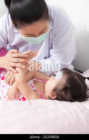 Ärztin überprüft und berührt den Arm kleines asiatisches Kind, Mädchen liegt auf einem Krankenbett und kühlende Fieberflecke auf ihrer Stirn. Studioaufnahme Stockfoto