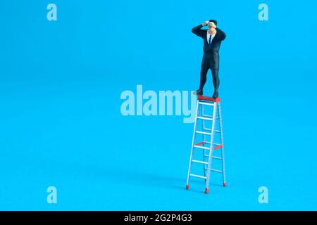 Geschäftsmann, der ein Fernglas über einer Klappleiter verwendet. Miniatur winzigen Menschen Spielzeug Fotografie. Isoliert auf blauem Hintergrund. Stockfoto