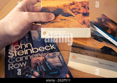 Eine Männerhand, die ein Taschenbuch der Trauben des Zorns hält, ein Roman des amerikanischen Autors John Steinbeck, mit anderen Romanen im Hintergrund Stockfoto