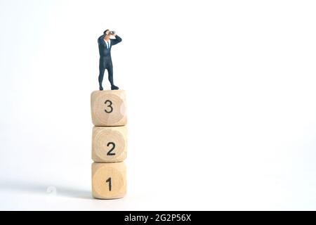 Miniatur Menschen Spielzeug Fotografie. Ein Geschäftsmann schaut, sucht, beobachtet mit einem Fernglas über den Würfeln isoliert auf weißem Hintergrund. Stockfoto