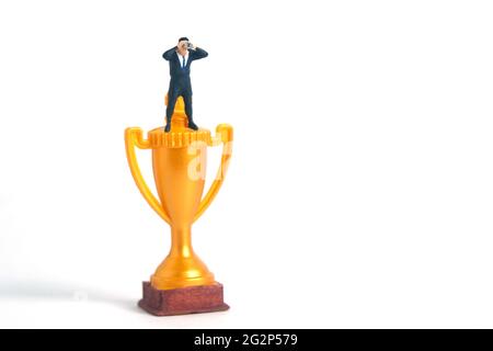Miniatur Menschen Spielzeug Fotografie. Ein Geschäftsmann schaut, sucht, beobachtet mit einem Fernglas über der goldenen Trophäe auf weißem Hintergrund. Stockfoto