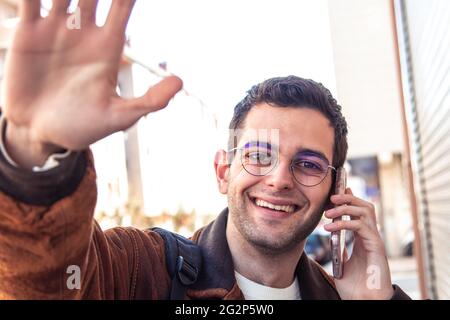 Porträt eines jungen Mannes, der lächelt und mit dem Mobiltelefon auf der Straße winkt Stockfoto