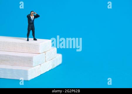 Geschäftsleute, die mit einem Fernglas über einer weißen Treppe stehen. Miniatur winzigen Menschen Spielzeug Fotografie. Isoliert auf blauem Hintergrund. Stockfoto