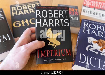 Eine Männerhand, die ein Taschenbuch von Imperium, einem historischen Roman des englischen Autors Robert Harris, mit anderen Romanen im Hintergrund hält Stockfoto