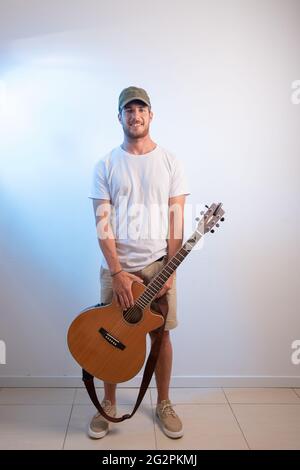 Ein attraktiver Musiker posiert mit seiner Akustikgitarre, er ist ein junger Künstler, er trägt eine Mütze und ein weißes Hemd. Person auf weißem Hintergrund isoliert. Stockfoto