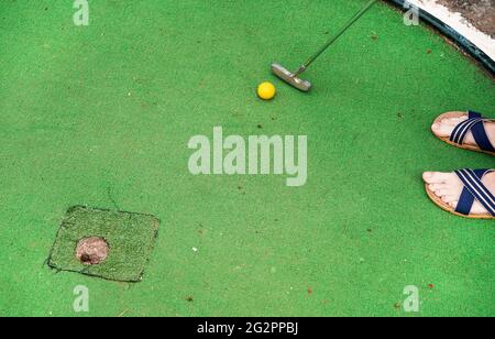 Junger Mann spielt Minigolf, Nahaufnahme von oben auf seinen Füßen in Sandalen, gelber Kugel, Metallknüppel und abgenutztem grünen Boden in der Nähe des Lochs Stockfoto