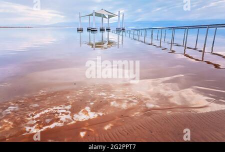 Ruhiger Morgen am Strand am Toten Meer von ein Bokek, rosa Wolken spiegeln sich über der Wasseroberfläche in der Nähe von Stahlschienen, die zum Sonnenschutz führen, Salzkristalle auf uns Stockfoto