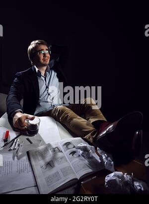 Ein Mann mittleren Alters in legerer Kleidung und Brille sitzt mit seinen Füßen auf dem Tisch mit Büchern und Papieren und trinkt Tee Stockfoto