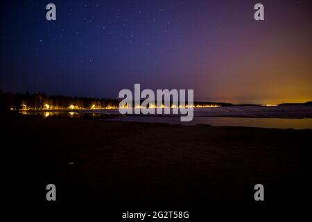 Sternenklare Nachtlandschaft vor einem halb zugefrorenen See in der Nacht in schweden mit orangefarbenen Horizont-Lichtern umgeben von Bäumen Stockfoto