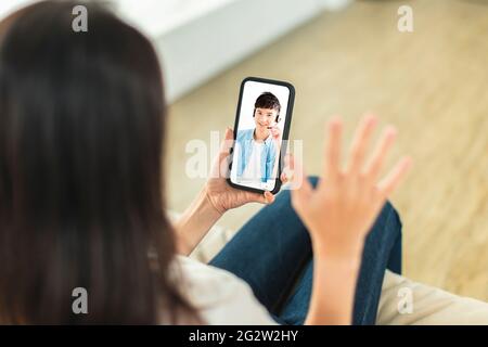 Junge Frau, die zu Hause auf dem Sofa sitzt, das Mobiltelefon hält, Videoanruf macht, mit Kollegen des Unternehmens über Telearbeit diskutiert Stockfoto