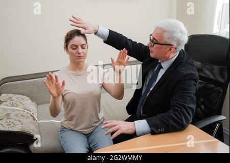 Reifer grauhaariger Mann hypnotisiert kaukasische Frau während der Hypnotherapie-Sitzung. Der Psychologe verwendet alternative Behandlungen für das Unterbewusstsein Stockfoto