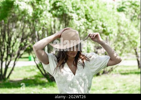 Junge schöne glückliche Frau in modischen Strohhut. Eine Brünette in einem weißen Hemd steht draußen und hält ihren Hut mit den Händen. Blühender Frühling A Stockfoto