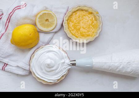 Herstellung von Mini-Zitronen-Baiser-Pasteten in Tartlet-Formen, eine mit, eine ohne Baiser, und Zitronenquark, und einem Paspelierbeutel voller Baiser Stockfoto