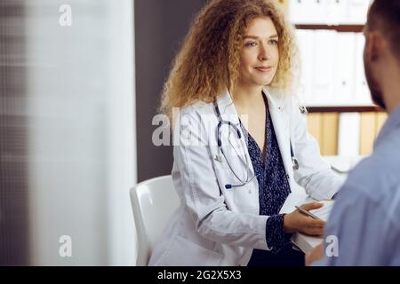 Ärztin und Patientin besprechen die aktuelle Gesundheitsuntersuchung, während sie in der sonnigen Klinik sitzen. Medizinkonzept Stockfoto