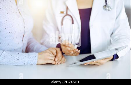 Unbekannte Ärztin zeigt ihrem Patienten eine Beschreibung der Medikamente, während sie zusammen am Schreibtisch in der sonnigen Vitrine in einer Klinik sitzt Stockfoto