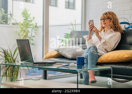 Frau zu Hause mit Handy und Computer auf dem Sofa sitzen Stockfoto