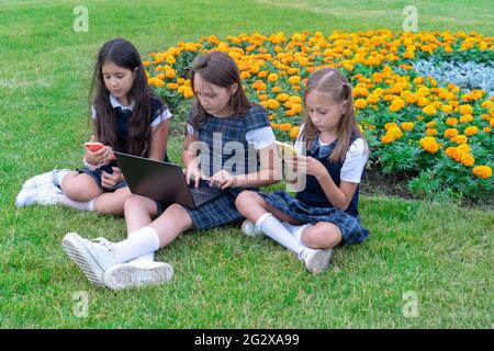 Drei Schülerinnen in Schuluniformen mit Telefon und Laptop sitzen an einem sonnigen Tag im Park auf dem Rasen. Fernunterricht und Outdoor-Trai Stockfoto