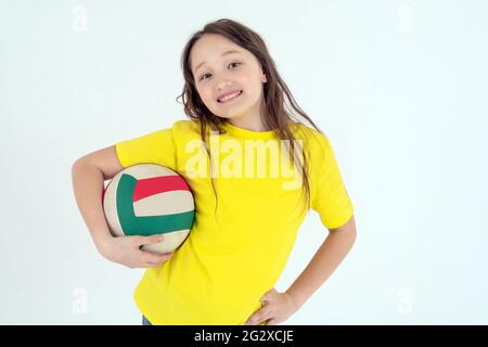 Ein Teenager-Mädchen in einem leuchtend gelben T-Shirt hält einen Volleyballball in der Hand und lächelt auf weißem Hintergrund. Studio-Foto. Stockfoto